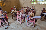 Djeca sjede za stolovima u improviziranoj učionici na čijem je podu pijesak