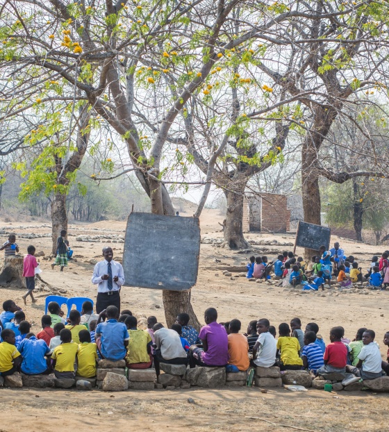 Djeca prate nastavu sjedeći na kamenju ispred drveta na kojem je ploča